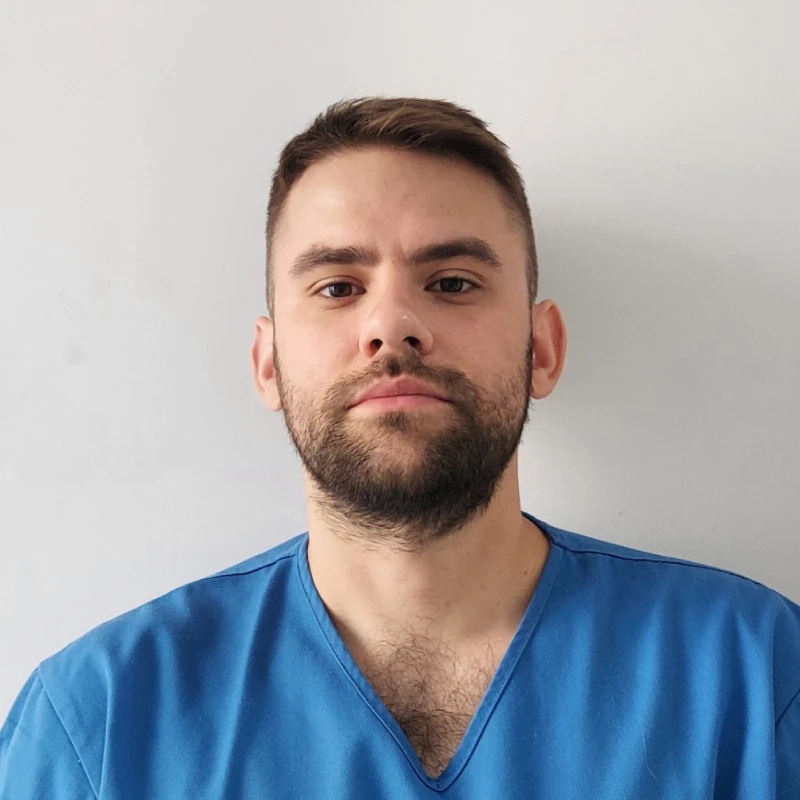 Muškarac u medicinskoj uniformi ispred bele pozadine, gleda pravo u kameru.