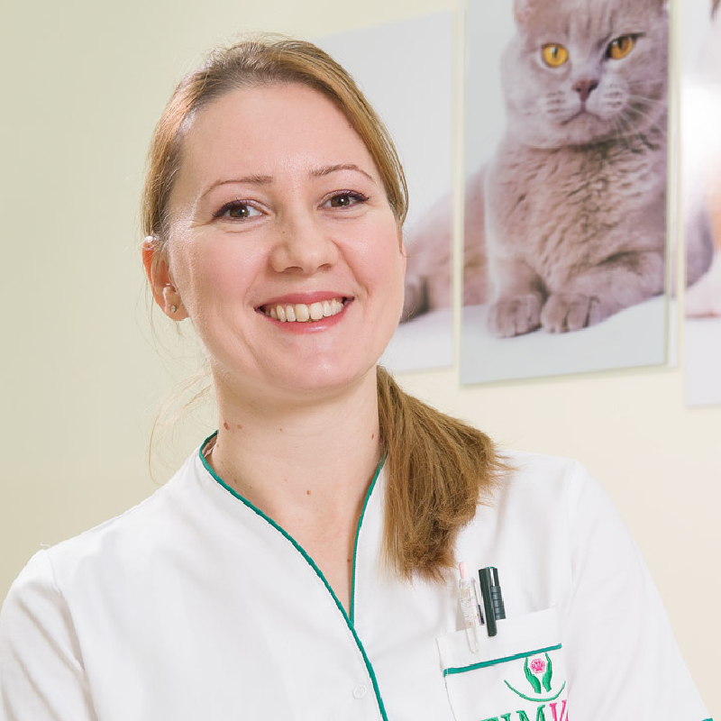 Veterinarka u belom mantilu sa osmehom, stoji ispred postera mačke u veterinarskoj ordinaciji.